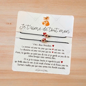 Puede incluir: Dos pulseras de amistad negras con cuentas de corazón rojas y una tarjeta blanca con el texto "Je t'aime de tout mon" y un mensaje sobre las pulseras. La tarjeta presenta una ilustración de dibujos animados de un zorro y un cachorro de zorro.