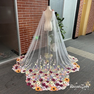 Puede incluir: Una capa de novia de tul blanco con un diseño floral. La capa está decorada con flores bordadas en tonos de rosa, morado, amarillo y naranja. La capa se muestra en un maniquí blanco.