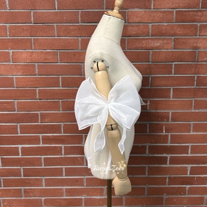 May include: A white sheer fabric bow tied around a mannequin's arm. The bow is large and fluffy, with a soft, delicate texture.