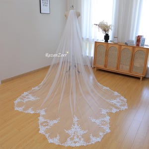 Puede incluir: Un velo de novia blanco y largo con un delicado ribete de encaje en el borde inferior. El velo se exhibe en un maniquí, mostrando su longitud y su intrincado diseño. El encaje presenta patrones florales, añadiendo un toque de elegancia.