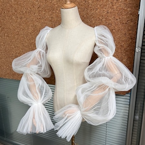 May include: White sheer fabric sleeves with multiple layers and a puffed design. The sleeves are made of a delicate, lightweight material and have a flowing, ethereal look.