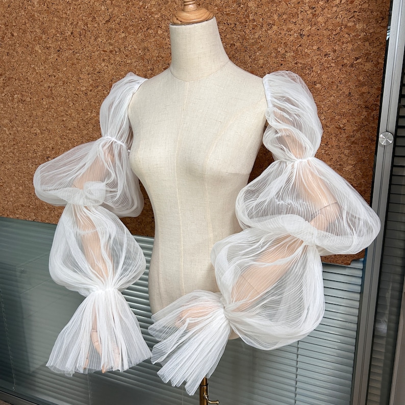 May include: White sheer fabric sleeves with multiple layers and a puffed design. The sleeves are made of a delicate, lightweight material and have a flowing, ethereal look.