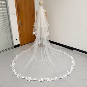 Puede incluir: Un velo de novia blanco de dos niveles con una larga cola. El velo se exhibe en un maniquí. Los bordes del velo están adornados con delicado encaje floral blanco. El velo está hecho de una tela transparente y ligera.