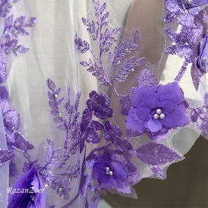 May include: Close-up of a sheer fabric adorned with intricate purple floral embroidery. The design features 3D flowers with pearl-like centers and delicate leaf patterns. The fabric is a light, airy material, perfect for bridal or formal wear.
