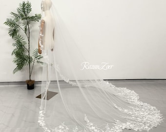 May include: A long, sheer white wedding veil featuring intricate floral lace embroidery along the edges. The veil is draped over a mannequin, extending to the floor with a detailed lace trim that showcases delicate leaf and petal patterns.