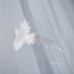 May include: A white tulle bridal veil featuring a delicate embroidered bird design. The bird is white with a gold shimmer and has its wings outstretched.