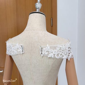 Può includere: Accessorio nuziale bianco in pizzo con spalle scoperte. Il delicato pizzo presenta motivi floreali e a foglie. L'accessorio è fissato con piccoli fermagli metallici. Il manichino è beige.