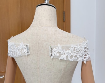 May include: White lace off-the-shoulder bridal accessory. The delicate lace features floral and leaf patterns. The accessory is attached with small metal clasps. The mannequin is beige.