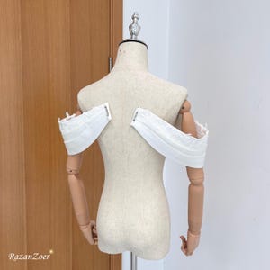 Puede incluir: Top blanco con hombros descubiertos y detalles de encaje, exhibido en un maniquí. El top presenta un diseño estructurado con una tela doblada y está diseñado para ser usado con los hombros descubiertos. El maniquí es beige y tiene brazos articulados.