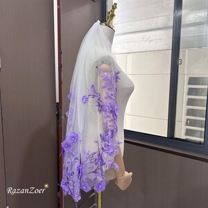 May include: A sheer white bridal veil with intricate purple floral embroidery and 3D flower accents. The veil is displayed on a mannequin, showcasing the delicate lace and design. The text "RazanZoer" is visible in the bottom left corner.