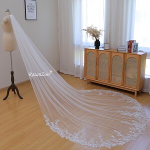 May include: A long, white bridal veil with a delicate lace trim. The veil is draped over a mannequin and is laying on a wooden floor.