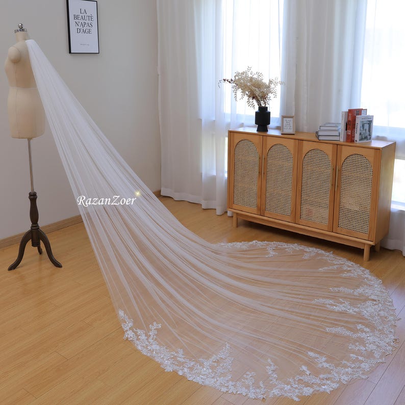 May include: A long, white bridal veil with a delicate lace trim. The veil is draped over a mannequin and is laying on a wooden floor.