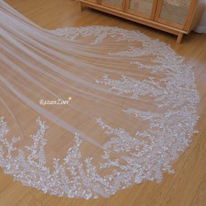 Puede incluir: Un velo de novia de tul blanco con un delicado ribete de encaje floral. El encaje presenta un patrón de hojas con pequeños adornos brillantes.