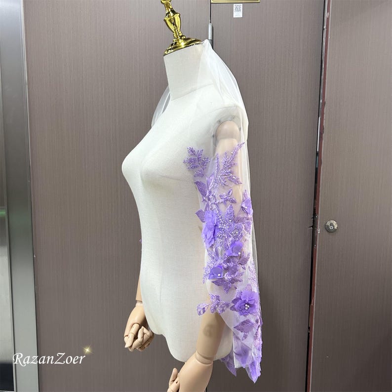 May include: A sheer white bridal veil with intricate lavender floral embroidery cascading down the sleeve. The delicate design features 3D flowers and beaded accents, creating a romantic and elegant look. The veil is displayed on a mannequin.