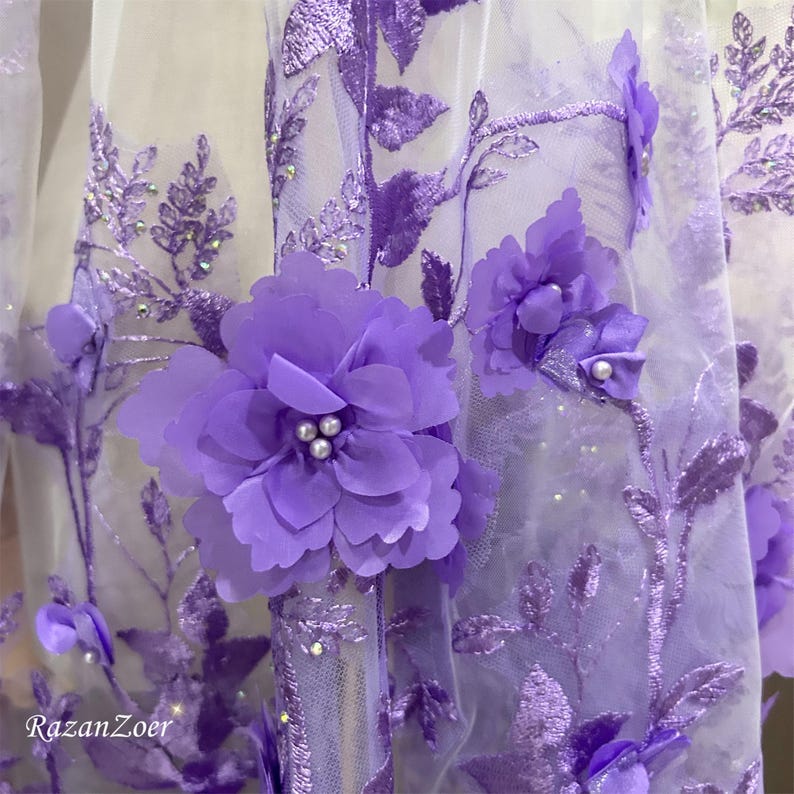 May include: Close-up of a sheer fabric with intricate floral embroidery. The design features large, three-dimensional purple flowers with pearl-like centers, and delicate embroidered vines and leaves. The fabric is a soft white, with the brand name "RazanZoer" visible.
