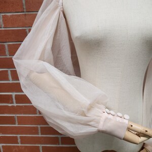 May include: A sheer, light pink blouse with long, puffy sleeves and white button detailing at the cuffs.