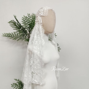 May include: A white lace bridal veil featuring intricate floral patterns and scalloped edges, adorned with small pearl accents. The veil is draped over a mannequin head and shoulders, with the text RazanZoer visible in the lower right corner.