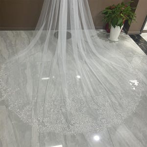 May include: A white tulle wedding veil with a beaded trim. The veil is long and flowing, and the beads are scattered throughout the trim.