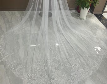 May include: A long, white, sheer tulle wedding veil featuring an intricate, scalloped lace border adorned with shimmering silver sequins and beads that extend along the curved hemline, creating a delicate and ornate decorative pattern.