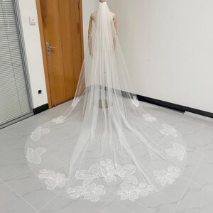 May include: A long, white bridal veil with floral lace appliqués. The veil is displayed on a mannequin, showcasing the delicate, sheer fabric and intricate embroidered details. The veil's design features a cascade of floral patterns.