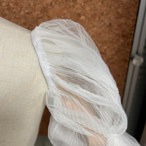 May include: Close-up of a white tulle fabric with a gathered, puffed sleeve detail. The fabric is soft and delicate, with a slightly sheer texture.