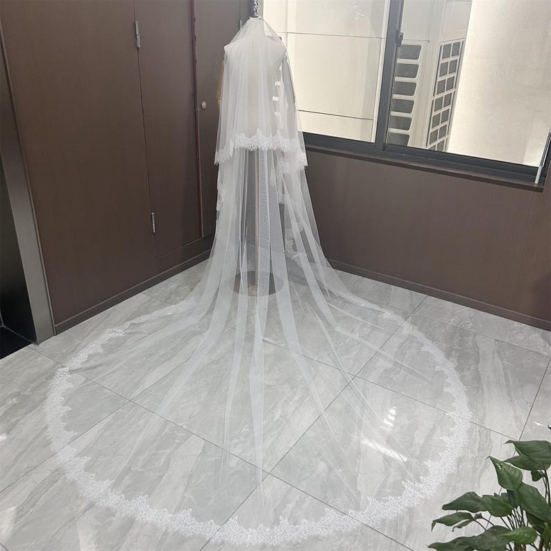 May include: A long, white bridal veil with a delicate lace trim. The veil is displayed on a mannequin, showcasing its length and intricate design. The veil's sheer fabric and detailed edging create an elegant and romantic look.