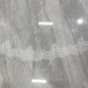 May include: A white bridal veil with a delicate lace trim. The lace features a floral pattern along the edge. The veil is made of a sheer, lightweight fabric, and is displayed against a gray marble surface.