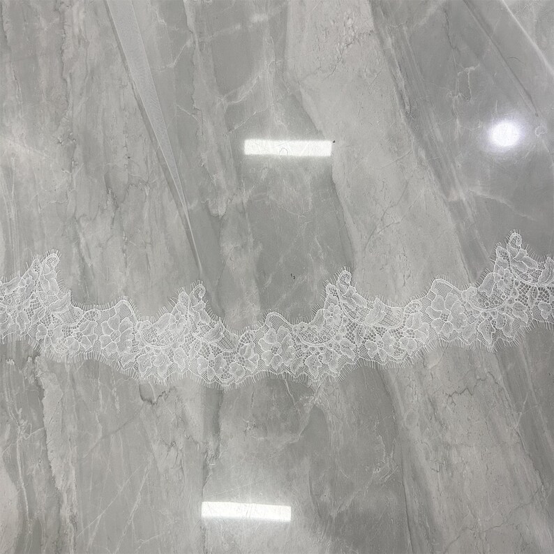 May include: A white bridal veil with a delicate lace trim. The lace features a floral pattern along the edge. The veil is made of a sheer, lightweight fabric, and is displayed against a gray marble surface.