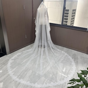 May include: A long, white bridal veil with a delicate lace trim. The veil is displayed on a mannequin, showcasing its flowing design and intricate details. The veil's length extends across the floor, highlighting its elegance.