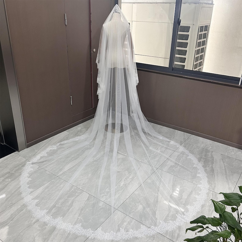 May include: A long, white bridal veil with a delicate lace trim. The veil is displayed on a mannequin, showcasing its flowing design and intricate details. The veil's length extends across the floor, highlighting its elegance.