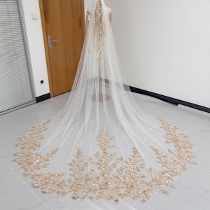 May include: A long, white bridal veil with detailed gold floral embroidery along the edges and a central design. The veil is displayed on a mannequin, highlighting its length and delicate features. The gold embellishments add a touch of elegance and sophistication.