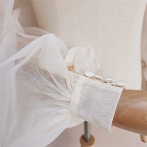 May include: Close-up of a white sheer blouse with puffed sleeves and white button detailing.