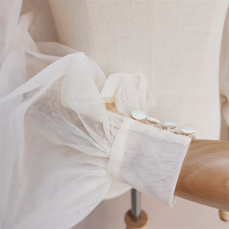 May include: Close-up of a white sheer blouse with puffed sleeves and white button detailing.