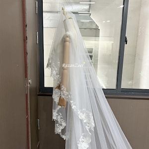 May include: A white bridal veil with a single layer of delicate lace trim. The veil is draped over a mannequin and is shown against a window.