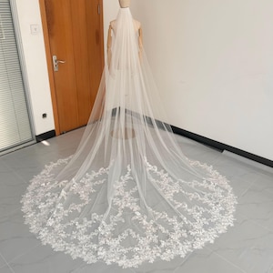 May include: A long, white bridal veil with detailed floral lace edging. The veil is displayed on a mannequin, highlighting its length and delicate design. The veil is made of a sheer, lightweight fabric, perfect for a wedding.