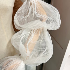 May include: White sheer fabric with a ruffled texture. The fabric is gathered and bunched up, creating a voluminous and airy look.