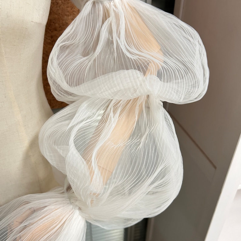 May include: White sheer fabric with a ruffled texture. The fabric is gathered and bunched up, creating a voluminous and airy look.