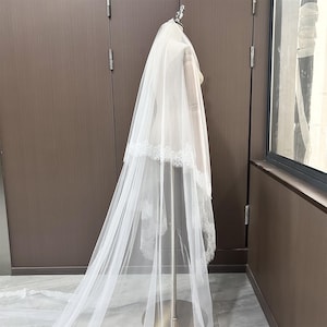 May include: A white bridal veil with a delicate lace trim. The veil is displayed on a mannequin, showcasing its flowing, layered design. The veil is made of a sheer, lightweight fabric.