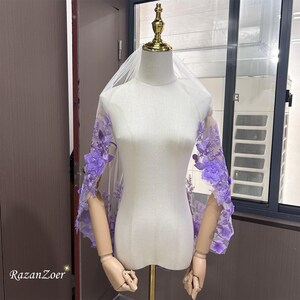 May include: A sheer white bridal veil with intricate lavender floral embroidery along the edges. The veil is draped over a mannequin, showcasing the delicate lace and three-dimensional flower details. The brand name "RazanZoer" is visible in the bottom left corner.