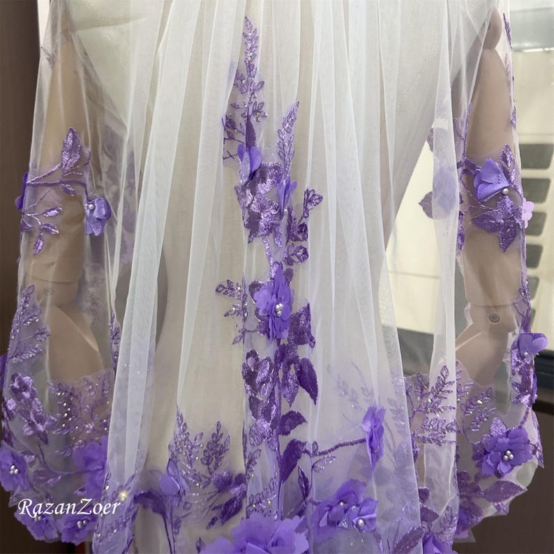 May include: A sheer white fabric with intricate purple floral embroidery and embellishments. The design features detailed purple flowers, leaves, and beaded accents, creating a delicate and elegant aesthetic. The text "RazanZoer" is visible in the bottom left corner.