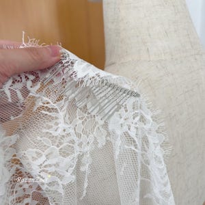 May include: Close-up of a white lace veil with a comb attached. The delicate lace features a floral pattern and is held up by a hand. The comb has silver metal teeth. The text "RazanZoer" is visible.