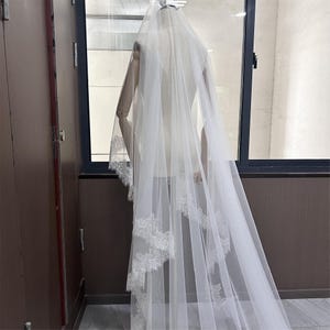May include: A long, white bridal veil with delicate lace trim. The veil is draped over a mannequin, showcasing its flowing design and intricate details. The lace is visible along the edges, adding a touch of elegance to the bridal accessory.