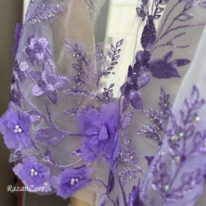 May include: Close-up of a sheer, lavender fabric with intricate floral embroidery. The design features 3D flowers, beaded accents, and delicate leaf patterns. The fabric is likely used for formal wear or special occasion garments.