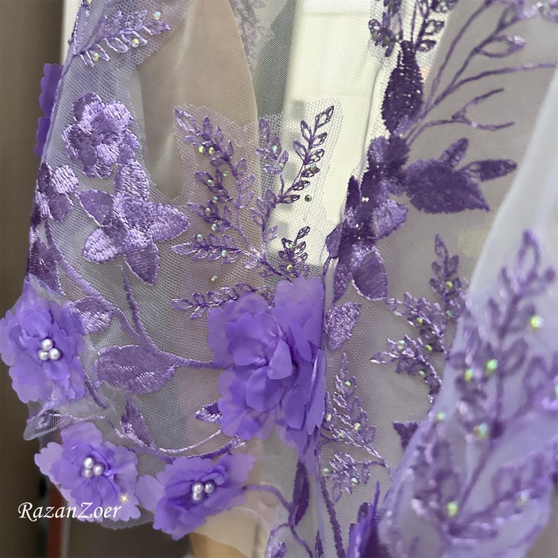 May include: Close-up of a sheer, lavender fabric with intricate floral embroidery. The design features 3D flowers, beaded accents, and delicate leaf patterns. The fabric is likely used for formal wear or special occasion garments.
