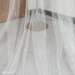 May include: A sheer white veil with a delicate pattern of sparkling stars and lines. The veil is draped, showcasing its flowing texture and subtle shimmer. The text "RazanZoer" is visible in the bottom left corner.