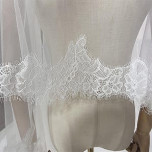 May include: A white bridal veil with delicate lace trim. The lace features a floral pattern and scalloped edges. The veil is made of a sheer, lightweight fabric, and is draped over a mannequin.