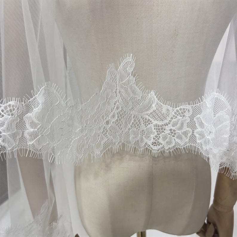 May include: A white bridal veil with delicate lace trim. The lace features a floral pattern and scalloped edges. The veil is made of a sheer, lightweight fabric, and is draped over a mannequin.