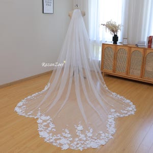 Puede incluir: Un velo de novia blanco con un borde de encaje floral. El velo es largo y fluido, con un delicado patrón de flores y hojas.