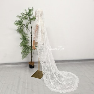 May include: A long white lace veil featuring intricate floral embroidery and scalloped edges, draped over a mannequin. The delicate fabric extends to the floor, showcasing a detailed pattern throughout the material with the text RazanZoer visible.