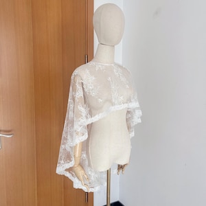 May include: A white lace cape with floral embroidery is displayed on a mannequin. The cape features a layered design with a scalloped edge, creating a delicate and elegant look. The sheer fabric allows the mannequin's form to be partially visible.